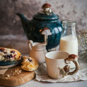 K&ouml;nnte beinhalten: Ein skurriles Stillleben mit einer tealfarbenen Teekanne und einer passenden Tasse mit Pilzverzierungen. Eine kleine Sahnek&auml;nnchen, eine Milchflasche und Geb&auml;ck erg&auml;nzen die Szene. Der Tassenhenkel ist ein Pilz, und die Teekanne hat einen Pilz auf dem Deckel.