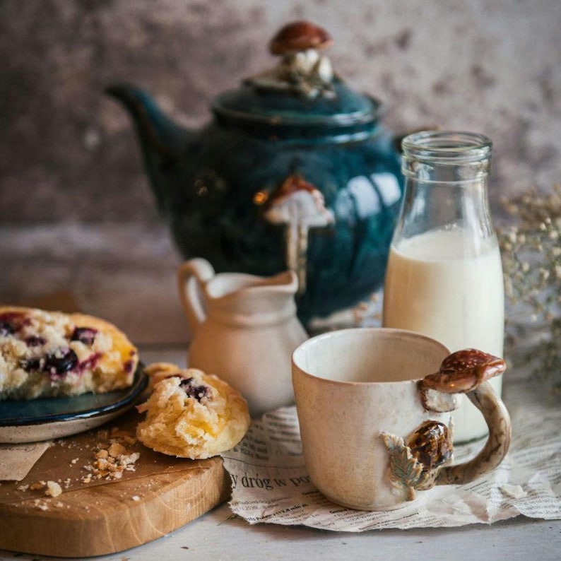 K&ouml;nnte beinhalten: Ein skurriles Stillleben mit einer tealfarbenen Teekanne und einer passenden Tasse mit Pilzverzierungen. Eine kleine Sahnek&auml;nnchen, eine Milchflasche und Geb&auml;ck erg&auml;nzen die Szene. Der Tassenhenkel ist ein Pilz, und die Teekanne hat einen Pilz auf dem Deckel.