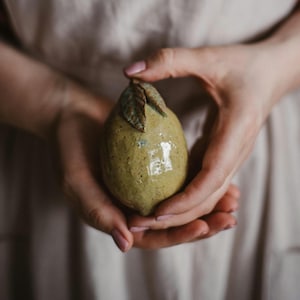 Könnte beinhalten: Eine Zitronenskulptur aus Keramik mit einer glänzenden, gesprenkelten gelb-grünen Glasur und einer strukturierten Oberfläche. Die Frucht hat ein kleines, braunes Blattdetail an der Oberseite und zeigt eine handgefertigte Optik.