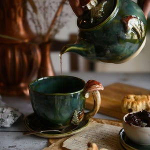 May include: A dark teal ceramic teacup and saucer set with a mushroom-shaped handle and decorative accents. A matching teapot pours tea into the cup. The set is on a table with a wooden cutting board and a small bowl of jam.