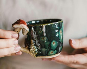 Könnte beinhalten: Eine handgefertigte Keramik-Tasse mit dunkelgrüner Glasur und einem einzigartigen Pilzgriff. Der Griff hat einen rot-weißen Pilzhut und einen detaillierten Stiel mit einem Blatt. Die Tasse wird von einer Person gehalten.