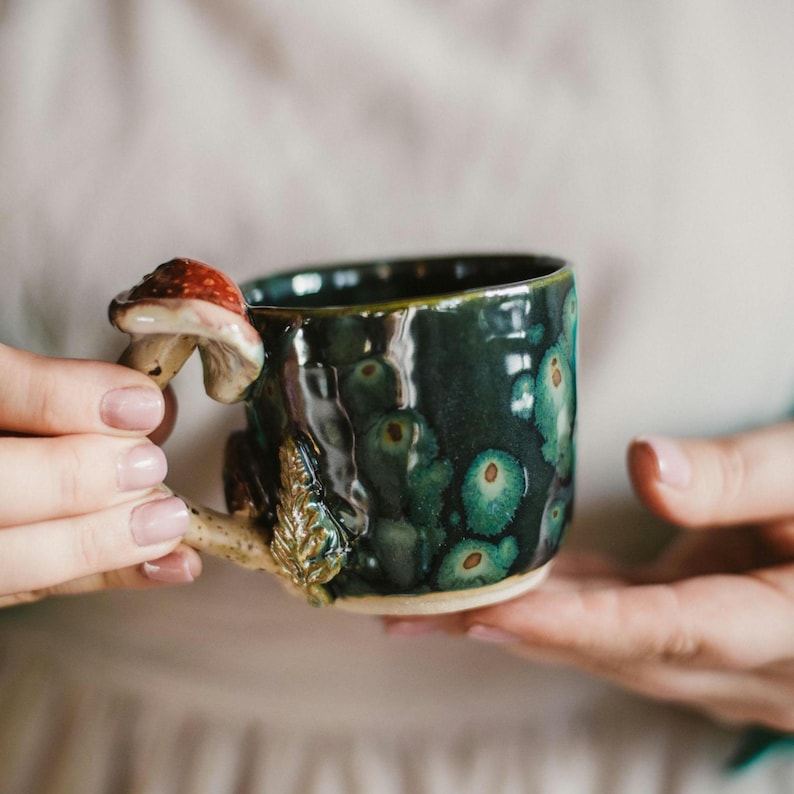 K&ouml;nnte beinhalten: Eine handgefertigte Keramik-Tasse mit dunkelgr&uuml;ner Glasur und einem einzigartigen Pilzgriff. Der Griff hat einen rot-wei&szlig;en Pilzhut und einen detaillierten Stiel mit einem Blatt. Die Tasse wird von einer Person gehalten.