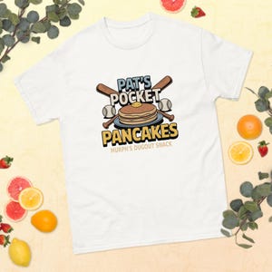 May include: White t-shirt featuring the text "PAT'S POCKET PANCAKES" with a stack of pancakes, baseballs, and crossed baseball bats. The phrase "MURPH'S DUGOUT SNACK" is below the pancakes. The shirt is surrounded by fruit and greenery.