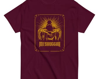Może przedstawiać: Bordowy t-shirt z złotym nadrukiem przedstawiającym postać w kapturze z rozłożonymi ramionami. Nadruk jest otoczony złotą ramką, a pod postacią widnieje napis "MESHUGGAH".