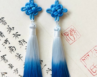 May include: A pair of blue and white tassel earrings. Each earring has a knotted blue top, a white base, and a blue ombre tassel. The earrings hang from silver hooks. The background features calligraphy and red stamps.