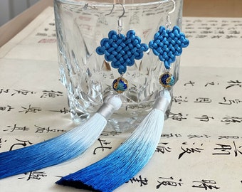 May include: A pair of blue and white tassel earrings with a knotted design and a blue and gold bead. The earrings are hanging from a glass cup on a table with a book.