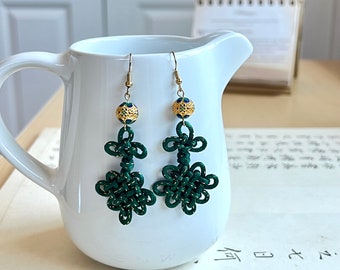 May include: A pair of green and gold earrings with a knot design. The earrings are hanging from a white surface.
