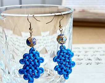 May include: A pair of blue and gold earrings with a knotted design. The earrings are made of a blue cord and have a small gold bead at the top.