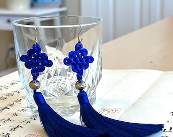 May include: A pair of blue tassel earrings with a knotted design and gold beads. The earrings are hanging from a glass.