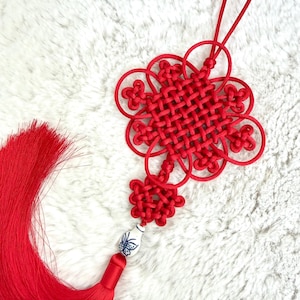 Può includere: Nodo cinese rosso, un ornamento decorativo, con intricati motivi intrecciati e un nappa. Il nodo ha un design centrale quadrato circondato da anelli a forma di fiore. Una piccola perla di ceramica bianca con un disegno di farfalla blu è attaccata alla nappa.