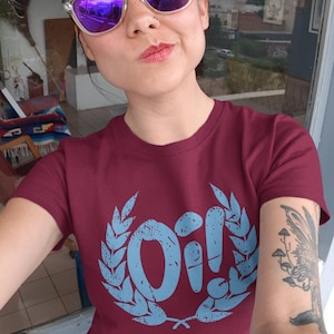 May include: A maroon t-shirt featuring a blue graphic of the word "Oi!" within a laurel wreath. The shirt is being worn by a person with a nose ring and purple sunglasses.