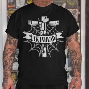 Op de afbeelding: Een zwart T-shirt met een witte afbeelding van een persoon op een kruis met een spinnenweb en de tekst "SKINHEAD".