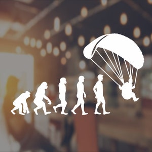 May include: A white silhouette graphic showing the evolution of paragliding. The image depicts a sequence of figures, evolving from ape-like forms to a person paragliding. The background is blurred, suggesting an outdoor scene.