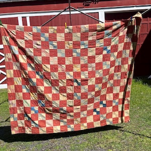 May include: A handmade quilt with a red and cream checkerboard pattern. The quilt is made of various fabric squares, some with floral and patterned designs. The quilt is hanging outside on a clothesline.