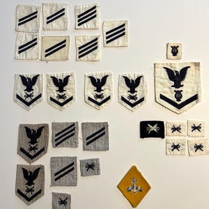 May include: A collection of vintage US Navy uniform patches, including rank insignia, specialty ratings, and a ship's anchor. The patches are made of white and blue fabric with embroidered details.