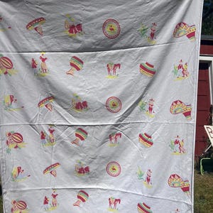 May include: White tablecloth with a repeating pattern of Mexican-themed illustrations. The design includes sombreros, pottery, figures, horses, and maracas in red, yellow, and green. The tablecloth is hanging outside on a clothesline.