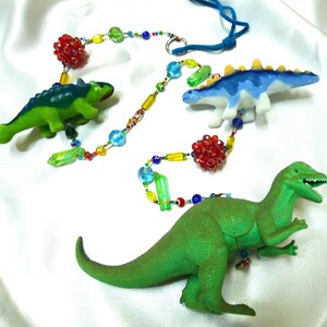 May include: A colorful beaded necklace with three dinosaur figurines. The necklace features red, yellow, blue, green, and clear beads. The dinosaurs are green, blue, and white.