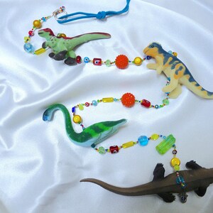 Puede incluir: Un collar de cuentas de colores con cuatro figuritas de dinosaurios. Los dinosaurios son verdes, azules, amarillos y marrones. Las cuentas son rojas, amarillas, azules, verdes y naranjas. El collar está enhebrado en un cordón azul.