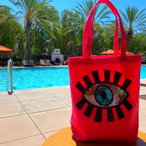 May include: A bright pink tote bag with a large, colorful eye design made of sequins. The eye is black with a blue and green iris. The bag is sitting on a brown surface near a swimming pool.