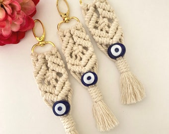 Puede incluir: Tres llaveros de macramé blanco con anillas doradas y dijes de ojo azul. Cada llavero tiene una borla en la parte inferior.