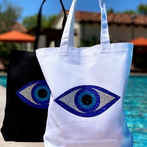 May include: Two canvas tote bags featuring a blue evil eye design. One bag is black and the other is white.