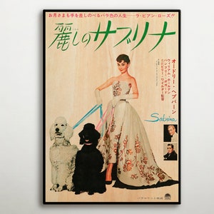 Op de afbeelding: Een vintage filmposter voor de film "Sabrina" met Audrey Hepburn, Humphrey Bogart en William Holden. De poster toont een kleurenbeeld van Audrey Hepburn in een witte jurk met een bloemenpatroon, die een riem vasthoudt waaraan twee poedels zijn bevestigd. De poster is in het Japans en bevat de titel "麗しのサブリナ" (Mooie Sabrina).