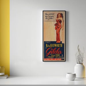 a framed vintage movie poster for the film "Gilda" by Rita Hayworth, hanging on a white wall. The poster has a yellow background and includes a picture of a woman in a red dress. There are also two small white vases and a small white bowl on a white surface below the poster.
