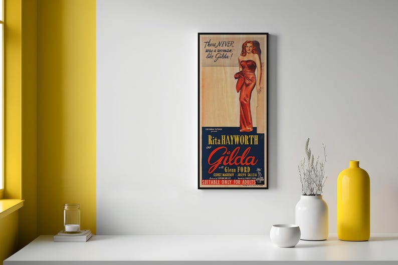 a framed vintage movie poster for the film "Gilda" by Rita Hayworth, hanging on a white wall. The poster has a yellow background and includes a picture of a woman in a red dress. There are also two small white vases and a small white bowl on a white surface below the poster.