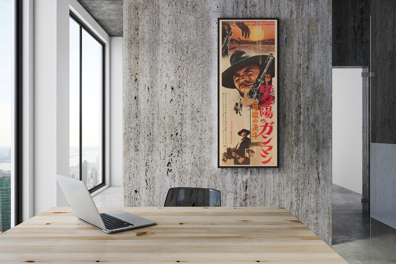 a wooden table with a laptop on it, situated in a room with a large window overlooking a cityscape. A framed poster of a man with a gun is hanging on the wall above the table.