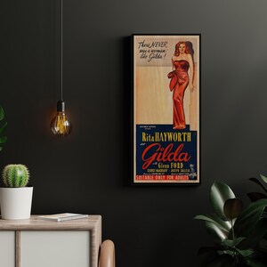 a framed vintage movie poster for the film "Gilda Gilda" by Rita Hayworth, with a woman in a red dress. The poster is hanging on a wall, and there are some plants and a lamp in the foreground.
