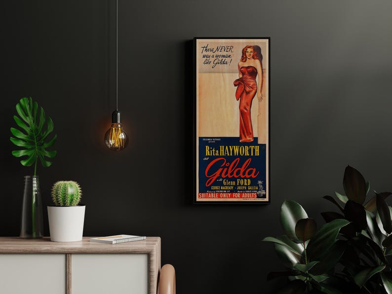 a framed vintage movie poster for the film "Gilda Gilda" by Rita Hayworth, with a woman in a red dress. The poster is hanging on a wall, and there are some plants and a lamp in the foreground.
