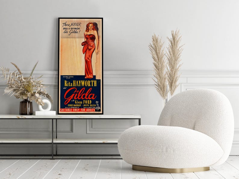 a framed vintage advertisement poster of Rita Hayworth and Gilda Gillett, with a white armchair and a potted plant in the foreground.