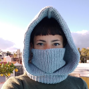 Puede incluir: Una combinación de capucha y cuello de punto azul claro. La capucha cubre la cabeza y el cuello cubre el cuello y la parte inferior de la cara. La persona de la imagen lleva un jersey verde oscuro. El fondo es un cielo azul claro.