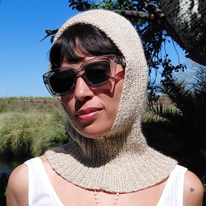 Puede incluir: Una capucha de punto beige y gafas de sol se usan con un vestido blanco. El vestido tiene un escote redondo y una cintura con adornos de encaje. El fondo presenta un río y árboles.