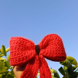 May include: A red knitted bow with a black center, held against a blue sky and green foliage.