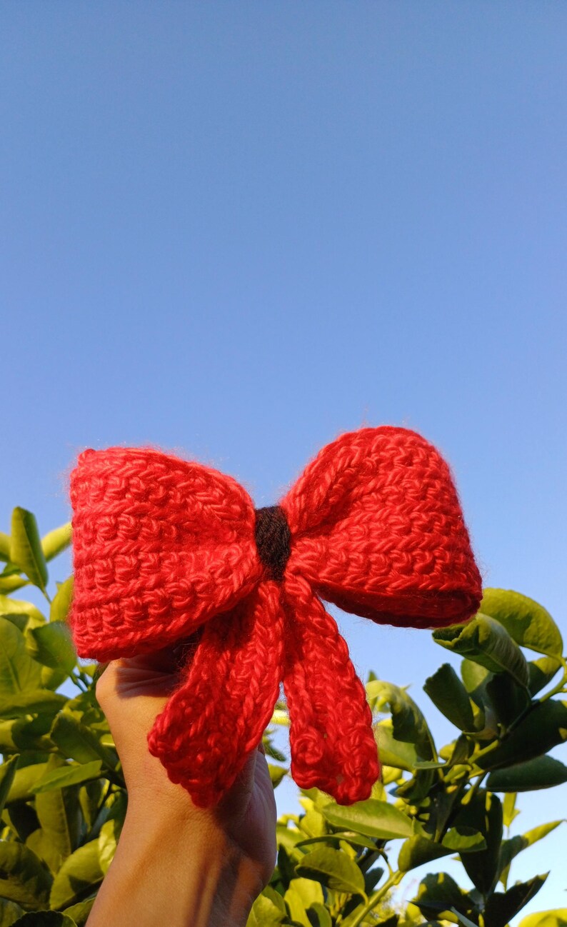 May include: A red knitted bow with a black center, held against a blue sky and green foliage.