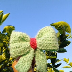 May include: A light green knitted bow with a pink center. The bow is held in a hand against a blue sky and green foliage background.