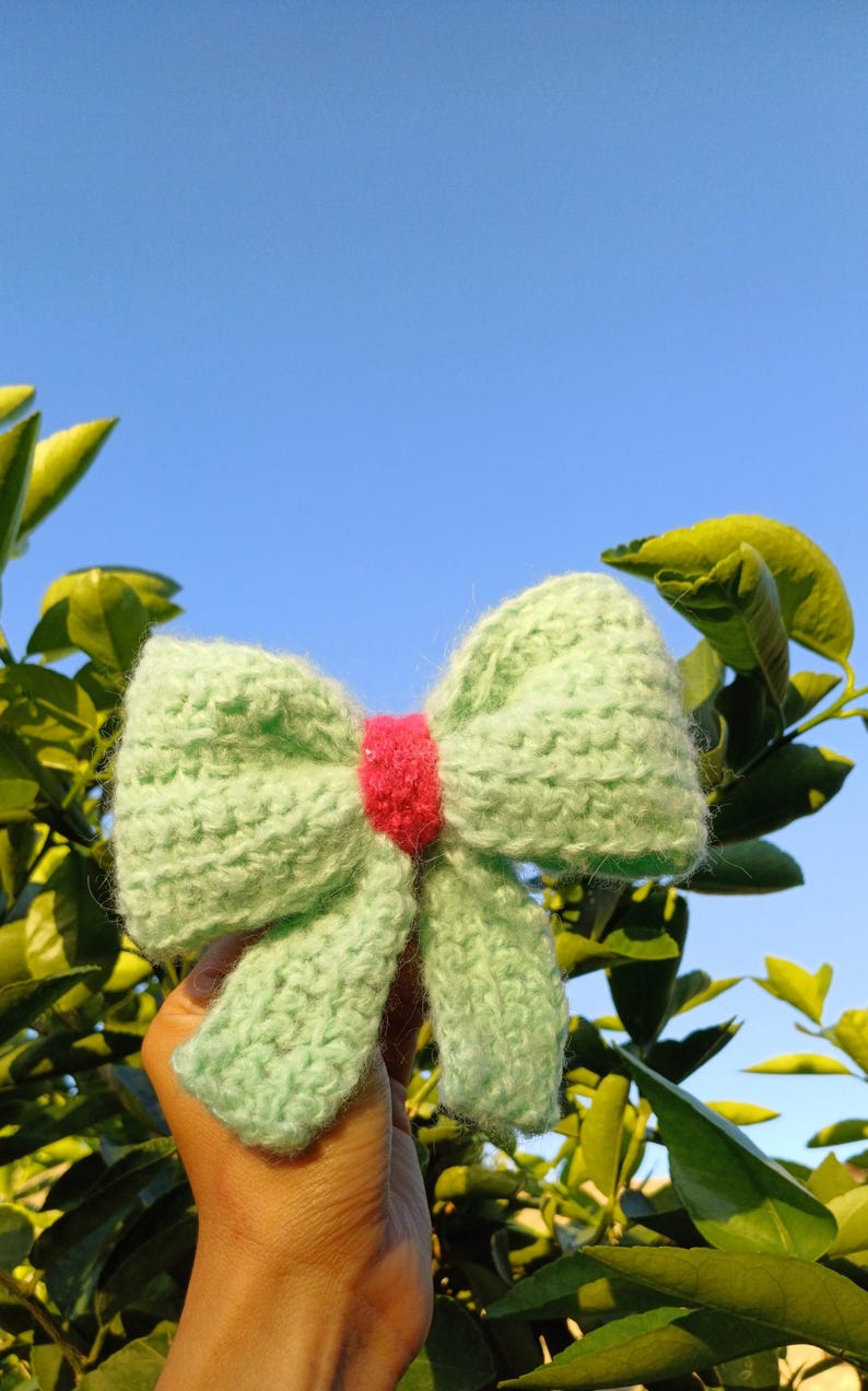 May include: A light green knitted bow with a pink center. The bow is held in a hand against a blue sky and green foliage background.