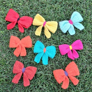 May include: A collection of ten crocheted bows in various colors, including red, orange, yellow, turquoise, and pink. The bows are arranged on a green grassy surface.