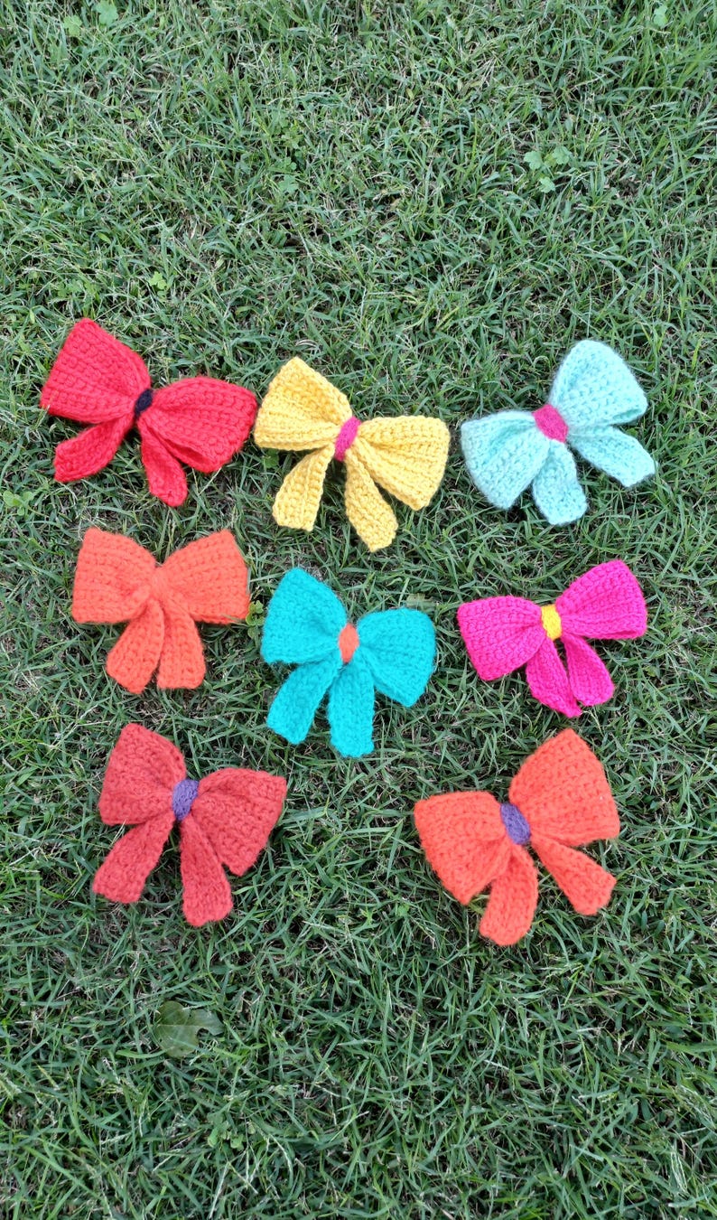May include: A collection of ten crocheted bows in various colors, including red, orange, yellow, turquoise, and pink. The bows are arranged on a green grassy surface.