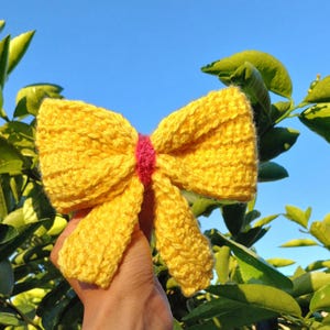 May include: A yellow crocheted bow with a pink center. The bow is held in a hand against a background of green leaves and a blue sky.