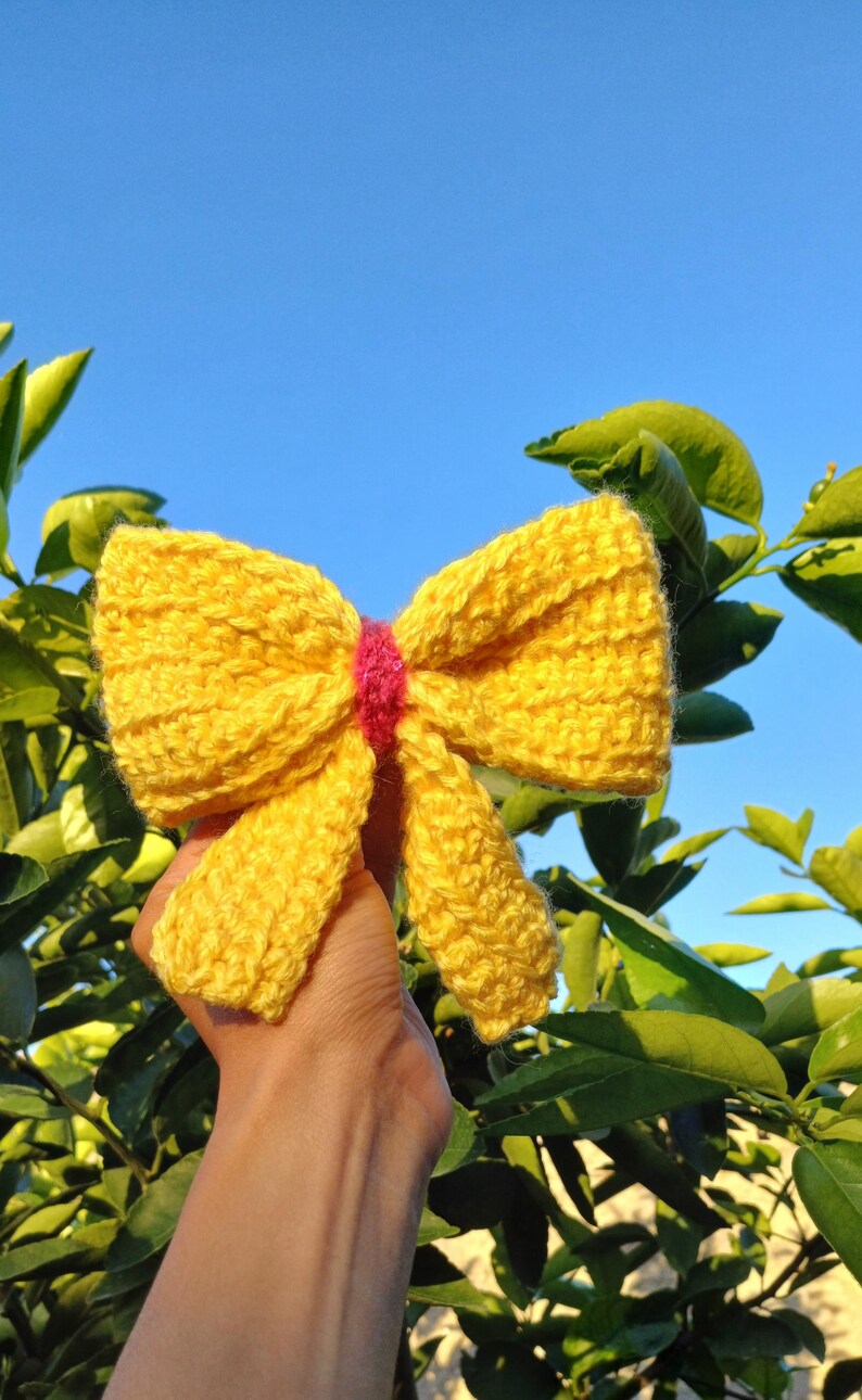 May include: A yellow crocheted bow with a pink center. The bow is held in a hand against a background of green leaves and a blue sky.