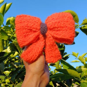 May include: A large, bright orange knitted bow with a small purple center. The bow is held in a hand against a background of green leaves and a blue sky.