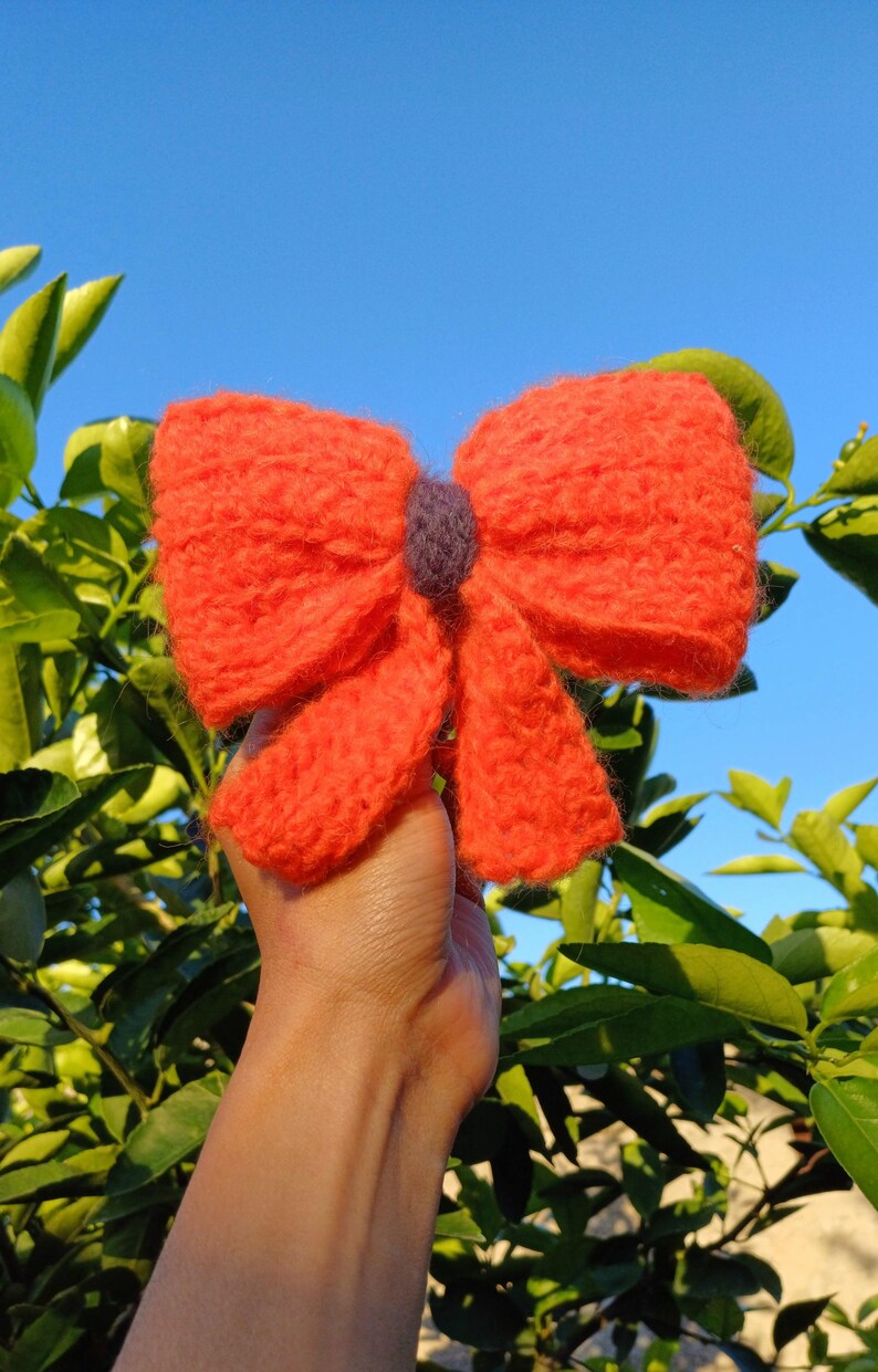 May include: A large, bright orange knitted bow with a small purple center. The bow is held in a hand against a background of green leaves and a blue sky.