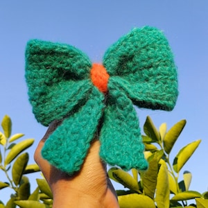May include: A teal blue knitted bow with a bright orange center. The bow is held up against a blue sky with green leaves in the background.