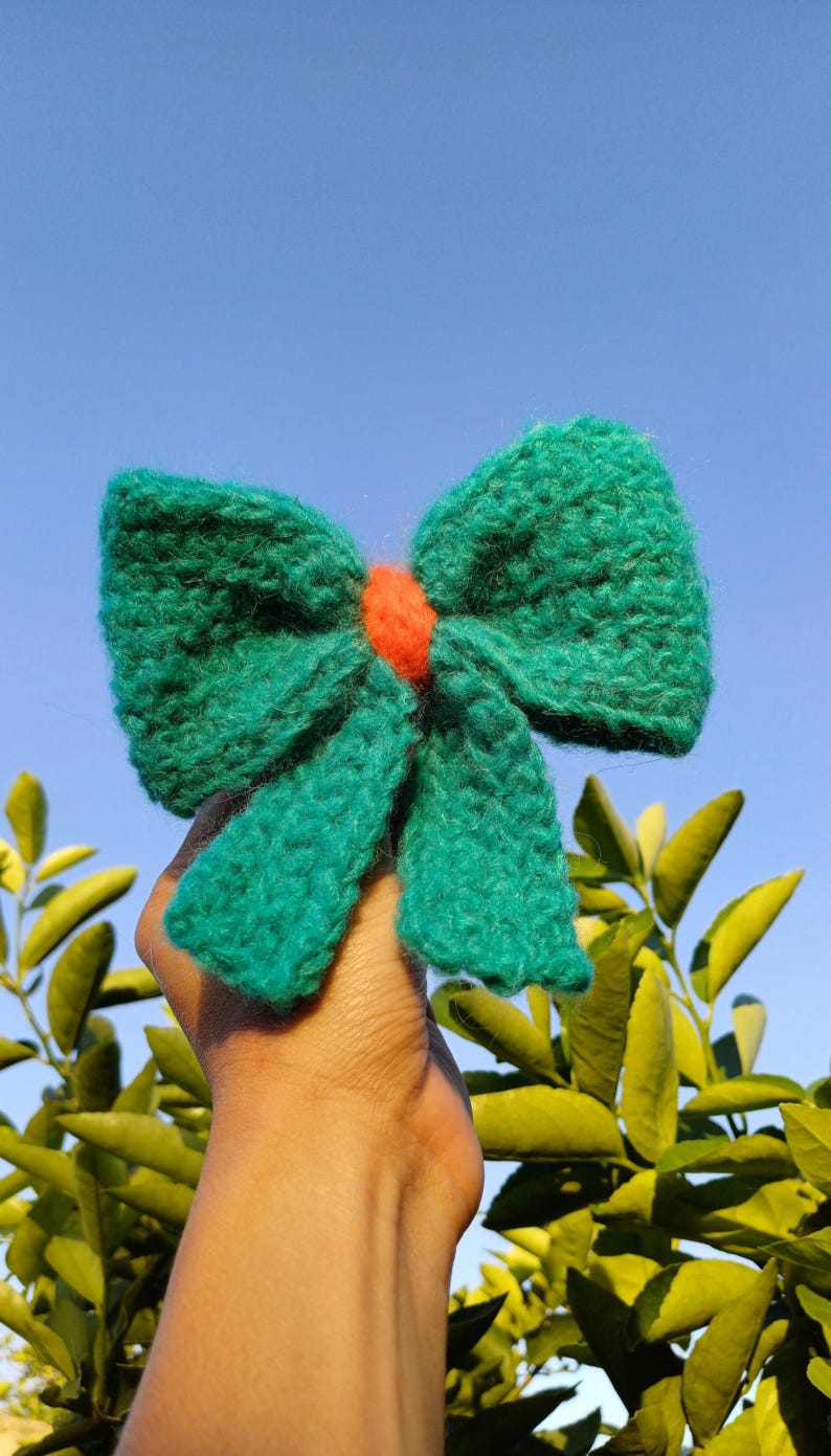 May include: A teal blue knitted bow with a bright orange center. The bow is held up against a blue sky with green leaves in the background.