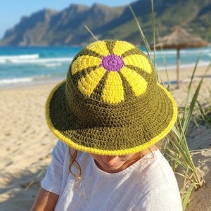 Puede incluir: Sombrero de sol de ganchillo con un gran diseño floral amarillo y morado en la corona. El sombrero es verde oliva con un borde amarillo. Se usa en un día soleado de playa.