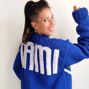 以下が含まれることがあります： 後ろに白い文字で「VAMI」と書かれた青いニットセーター。セーターの裾には緑と白のストライプが入っています。