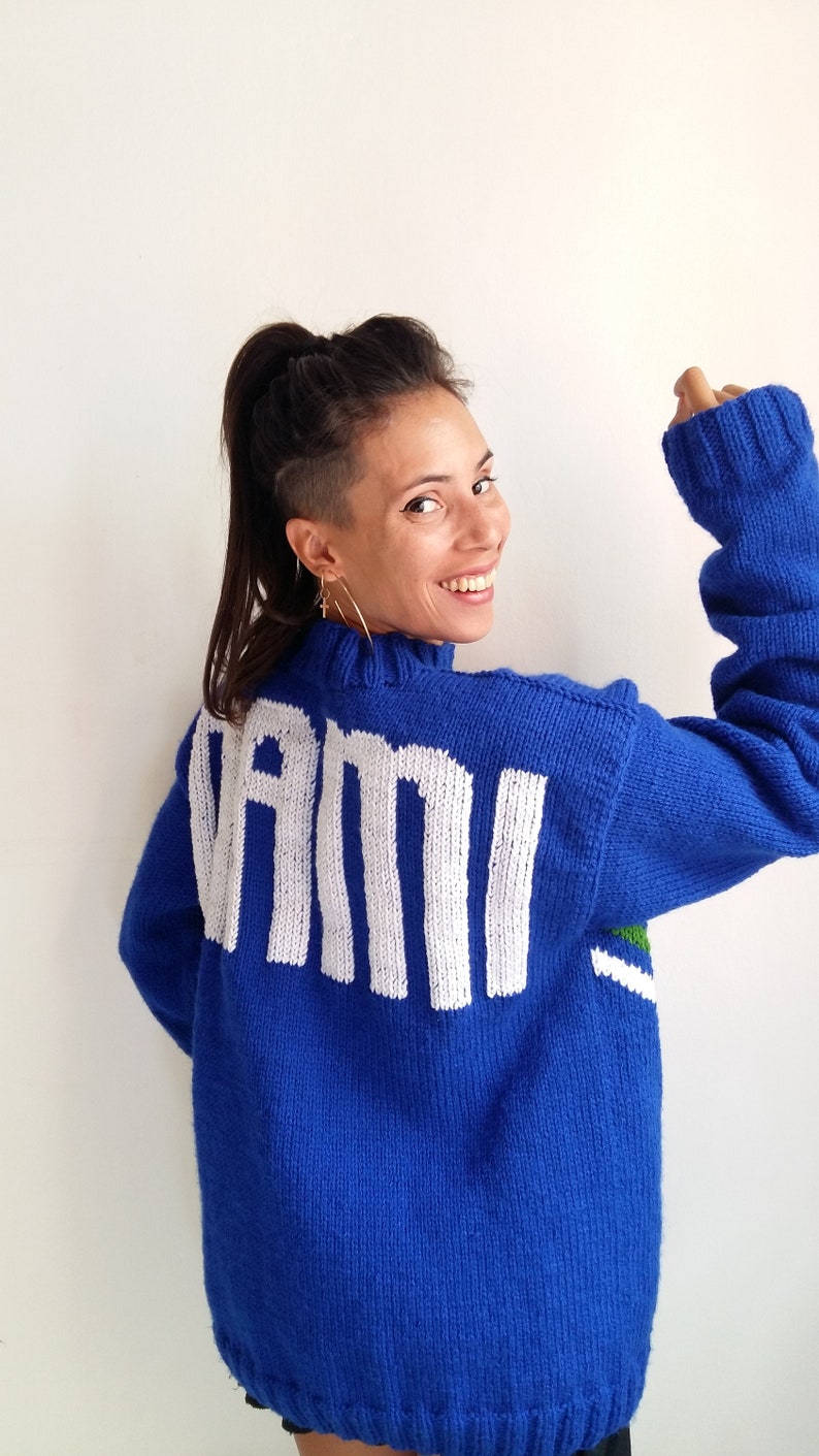 以下が含まれることがあります： 後ろに白い文字で「VAMI」と書かれた青いニットセーター。セーターの裾には緑と白のストライプが入っています。