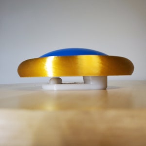 May include: A blue and gold button with a white base. The button is round and has a shiny gold rim.